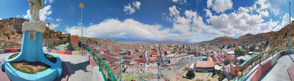 Panoramic cityscape of Oruro Bolivia showing colorful buildings in a valley surrounded by brown hills, with a white religious statue on a blue pedestal in the foreground