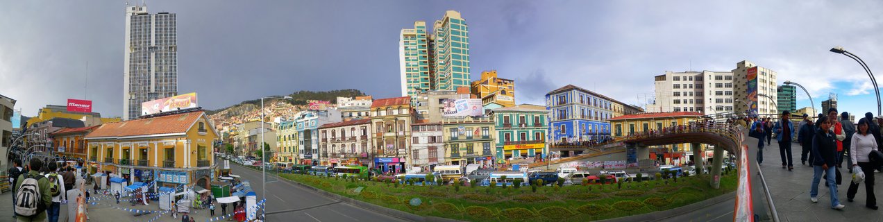 Wide panoramic street view of La Paz showing colorful colonial buildings, modern glass towers, pedestrians on walkways, vehicles on streets, and hillside housing under cloudy sky