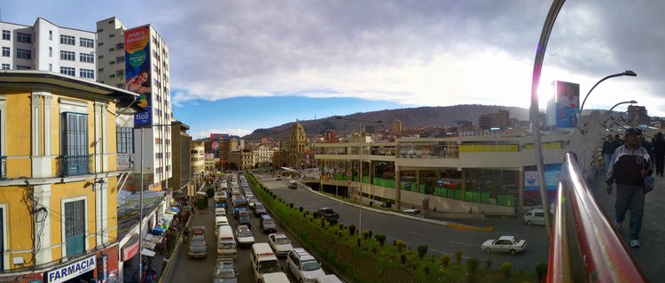 Panoramic view of a busy avenue in La Paz, Bolivia showing heavy traffic with white vans and buses, colonial yellow buildings, modern structures, a cathedral dome, street vendors, pedestrians on a walkway, and mountains in the background under cloudy skies
