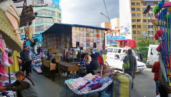 Outdoor street market in La Paz with vendors selling shoes, clothing, and household goods from stalls covered with blue tarps, surrounded by multi-story buildings and pedestrians
