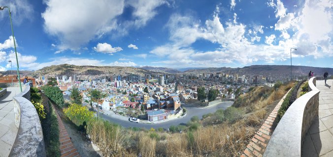 Panoramic cityscape of La Paz Bolivia showing densely packed buildings in a mountain valley with blue sky and white clouds above, viewed from an elevated stone walkway viewpoint