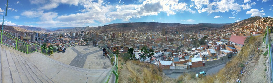 Panoramic cityscape of La Paz, Bolivia showing densely packed buildings covering a valley and hillsides, viewed from an elevated platform with green railings where people are seated on benches