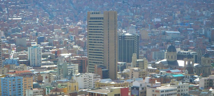 Aerial cityscape of La Paz, Bolivia showing dense urban development with a prominent beige high-rise tower, modern glass buildings, and colonial cathedral domes against a backdrop of tightly packed residential buildings