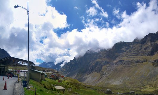 Mountain entrance station with covered checkpoint, traffic cones, and street light in foreground, with dramatic cloud-covered rocky peaks and green slopes in the Bolivian Andes