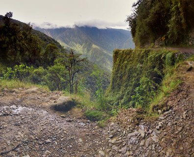 Rocky unpaved mountain road with steep moss-covered cliff on the right side, dropping into a deep green valley with forested mountains in the background under cloudy skies