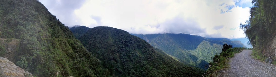 Panoramic view of a gravel road along a steep mountainside covered in dense green forest, with cloud-covered mountain peaks and a deep valley in the background near La Paz, Bolivia