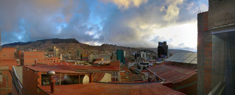 Panoramic cityscape of La Paz, Bolivia showing red rooftops in foreground, mid-rise and high-rise buildings climbing hillsides, with dramatic cloudy sky at golden hour