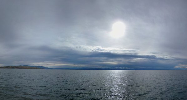 Wide view of Lake Titicaca with cloudy sky, sun breaking through clouds creating reflection on water, distant mountains visible on left horizon