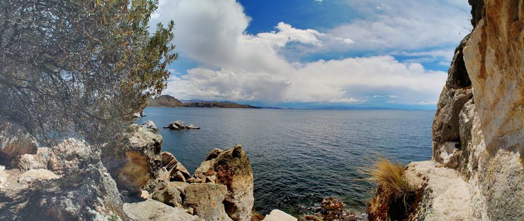 Panoramic view of Lake Titicaca showing rocky shoreline in foreground, deep blue water, distant islands, and cloudy sky