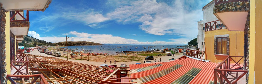 Panoramic view from a balcony overlooking Lake Titicaca in Copacabana, Bolivia, showing boats on blue water, colorful buildings with red roofs, and people on the shoreline under a partly cloudy sky