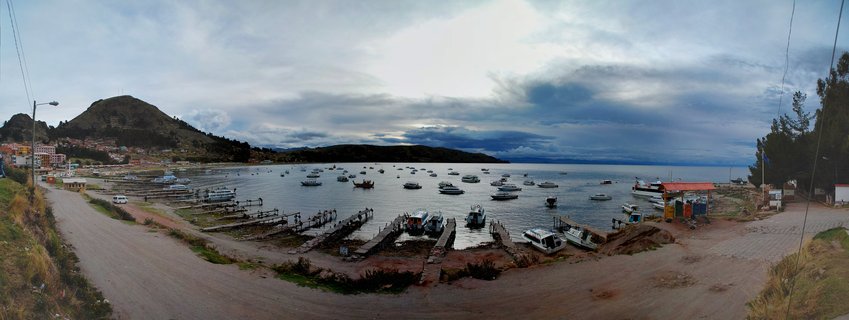 Panoramic harbor view showing boats anchored in Lake Titicaca with wooden docks, hills, and buildings under cloudy sky in Copacabana, Bolivia