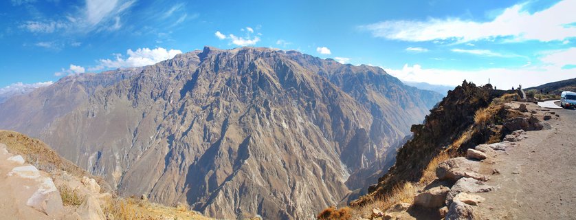 Panoramic landscape photograph showing Colca Canyon in Peru with steep mountain walls, blue sky with white clouds, and a viewing area with tourists and a bus on the right side