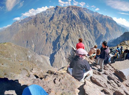 Group of tourists sitting and standing on rocky terrain at a viewpoint, with massive brown mountain canyon walls rising in the background under blue sky with white clouds