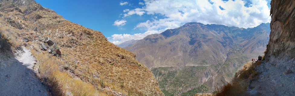 Mountain canyon landscape showing a dirt hiking trail on an arid slope, with steep valley walls, terraced green areas below, and distant mountain peaks under a blue sky with clouds
