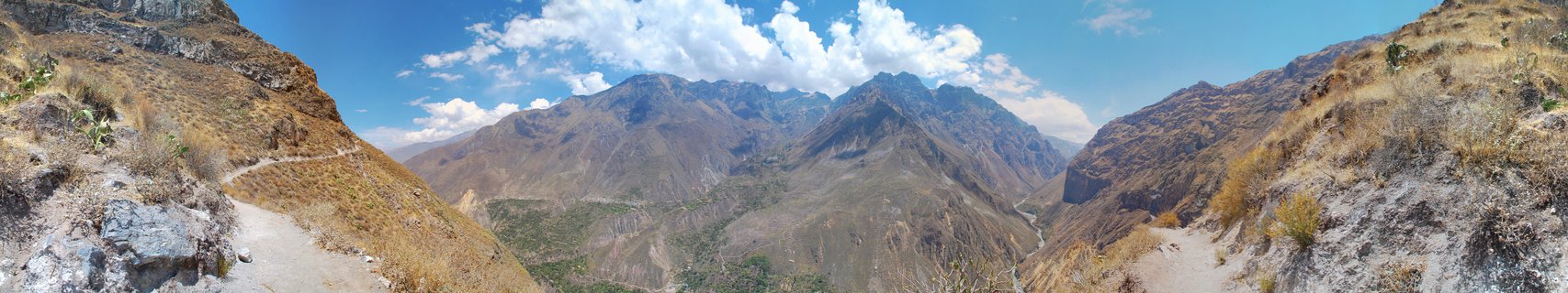 Panoramic view of Colca Canyon in Peru showing steep rocky mountain slopes with dry grass, a winding dirt trail, deep valley with green vegetation, and distant mountain peaks under blue sky with white clouds