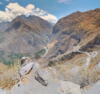 Wide landscape view of Colca Canyon in Peru showing steep brown mountain slopes, a river winding through the valley bottom, green vegetation patches, and a dirt road on the right side under blue sky with white clouds
