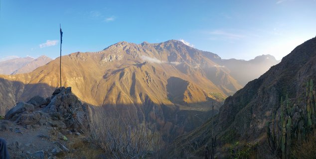 Mountain landscape showing golden-brown Andean peaks illuminated by morning sunlight under a clear blue sky, with rocky foreground, a tall pole marker, and cacti on the canyon slopes