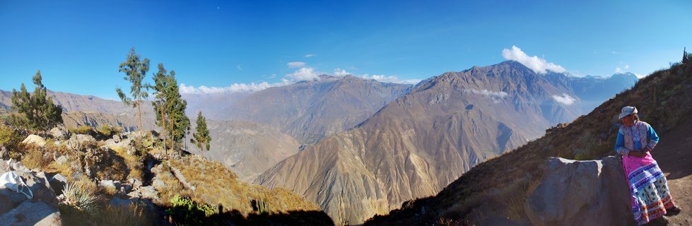 Panoramic view of Colca Canyon in Peru with steep mountain slopes, clear blue sky, and a woman in colorful traditional clothing standing at a viewpoint on the right side
