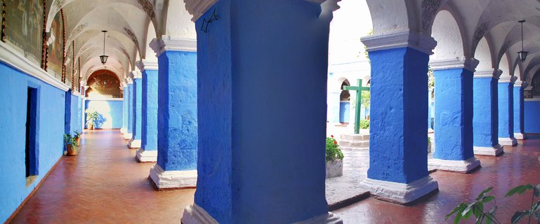 Covered walkway with bright blue columns and white arched ceilings surrounding an open courtyard with a green cross at a historic monastery in Peru