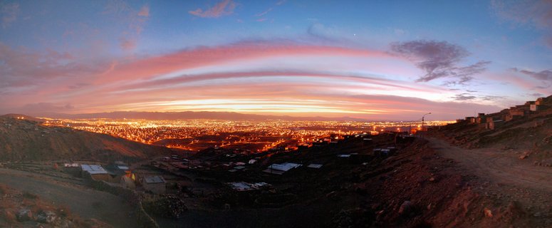 Panoramic cityscape of Cusco Peru at dusk showing illuminated city lights in a valley surrounded by dark mountains with pink and orange sunset clouds in a blue sky above