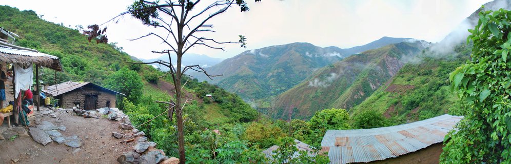 Panoramic mountain landscape showing green forested peaks with mist, rustic buildings with metal roofs, and a dirt path in the foreground near Machu Picchu, Peru