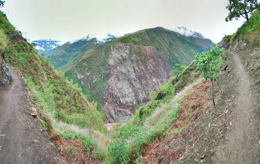 Mountain valley landscape showing two dirt hiking trails on green slopes with rocky cliff face and cloud-covered peaks in Machu Picchu, Peru