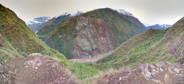 Panoramic mountain valley landscape at Machu Picchu, Peru, showing lush green cloud forest vegetation covering mountainsides, exposed gray cliff face with landslide scars, muddy brown Urubamba River in valley below, white clouds around mountain peaks, rocky foreground with dirt hiking trail, Andes mountain range in background under overcast sky