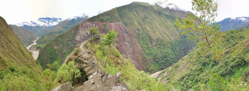 Mountain landscape showing green forested slopes, a winding river valley, stone pathway ruins, and clouds around mountain peaks in Peru