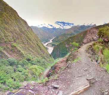 A panoramic view of a mountain hiking trail on a steep hillside overlooking a river valley with lush green mountains and clouds in the distance near Machu Picchu, Peru