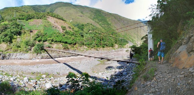 Hikers with large backpacks walking across a wooden suspension bridge over a rocky river with green mountains in the background in Peru