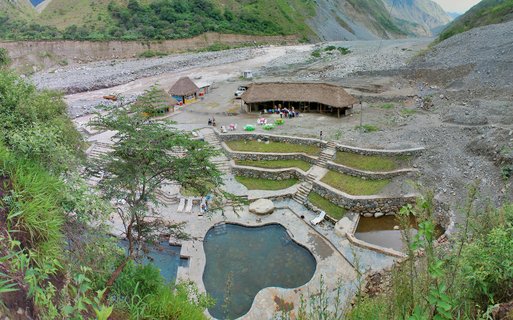 Aerial view of thermal hot springs with irregular blue pools, stone terraces with grass, thatched-roof buildings, and visitors in a mountain valley near Machu Picchu, Peru