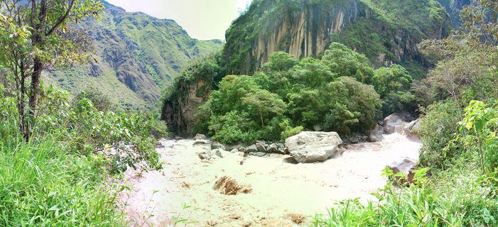 A muddy river flows through a mountain valley with steep green slopes and dense forest vegetation on both banks under overcast sky