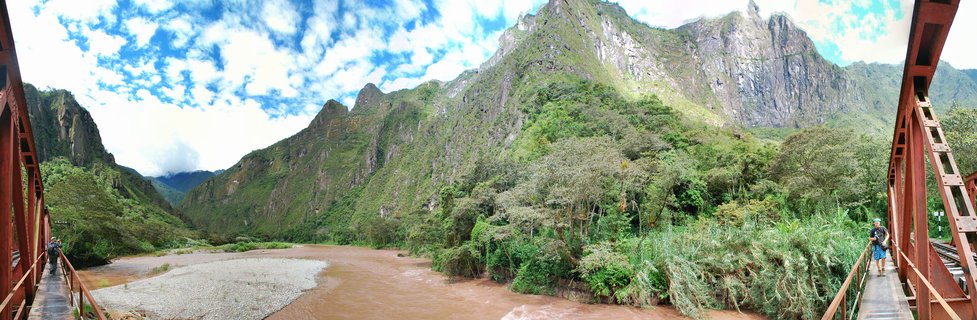 Panoramic landscape showing steep green mountains, a brown river, and red bridge structure with tourists, at Machu Picchu, Peru