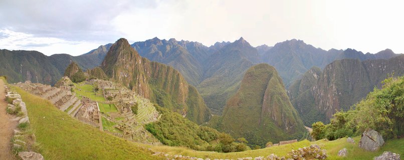Panoramic view of Machu Picchu archaeological site showing ancient Incan stone ruins and agricultural terraces on a mountainside, with Huayna Picchu peak rising in the background under an overcast sky in the Andes mountains of Peru