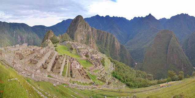 Aerial view of Machu Picchu archaeological site showing ancient Incan stone ruins and green terraces with Huayna Picchu mountain peak in the background, surrounded by forested Andean mountains under cloudy sky