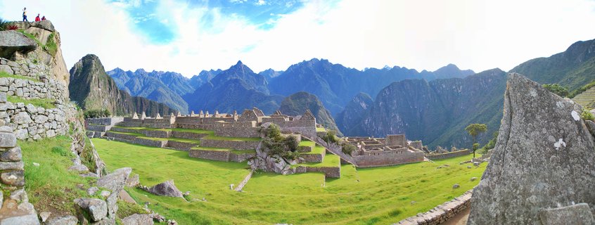 Panoramic view of ancient stone ruins of Machu Picchu with green terraced lawns, surrounded by steep mountain peaks under a blue sky with scattered clouds