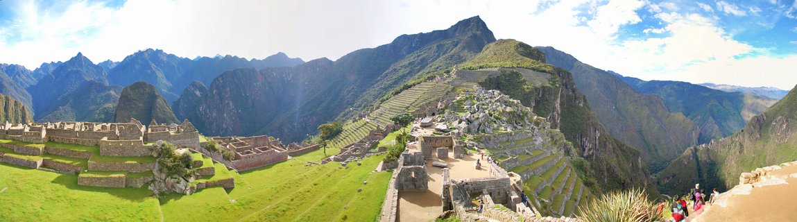 Panoramic photograph of Machu Picchu showing ancient stone Incan ruins with terraced structures, green grass, tourists walking on pathways, surrounded by steep mountain peaks under a blue sky with clouds