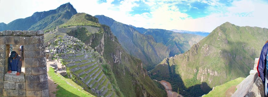 Panoramic view of Machu Picchu showing ancient stone ruins with terraced structures, Huayna Picchu mountain in background, green Andean peaks, and Urubamba River valley below under partly cloudy sky