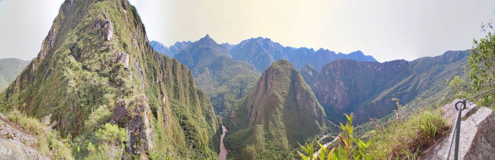 Panoramic landscape showing Machu Picchu mountain peaks covered in green vegetation with the Urubamba River valley below and distant Andean mountain ranges under a bright sky