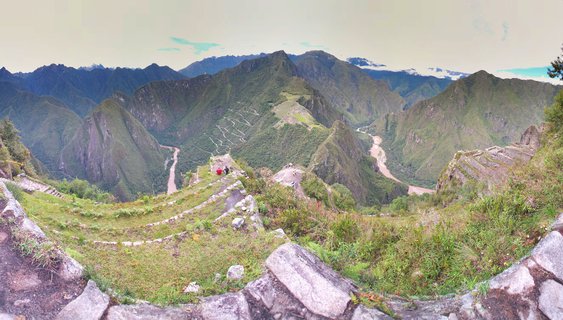Panoramic landscape showing ancient Incan stone ruins and terraces at Machu Picchu with green mountains, winding river, and cloudy sky in Peru