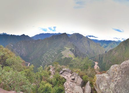 Panoramic view of Machu Picchu archaeological site with stone ruins, green mountains, Huayna Picchu peak, winding river, and cloudy sky in Peru
