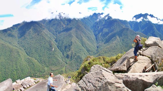 Tourists standing on ancient Incan stone ruins at Machu Picchu archaeological site with green mountain ranges and clouds in the background