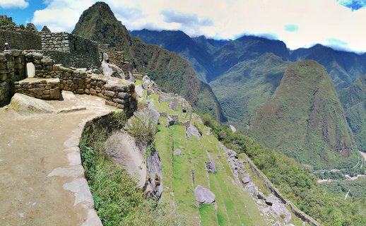 Ancient Incan stone ruins and agricultural terraces at Machu Picchu archaeological site in Peru, with Huayna Picchu mountain peak visible in the background under blue sky with white clouds. A small figure of a person stands on the stone pathways in the foreground.