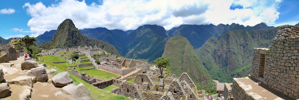 Panoramic photograph of Machu Picchu showing ancient Incan stone ruins with terraced fields, surrounded by green mountain peaks under a blue sky with white clouds