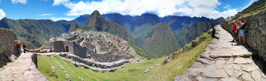 Panoramic photograph showing the ancient stone ruins of Machu Picchu with green terraced fields, stone pathways with tourists, and Huayna Picchu mountain peak rising in the background under a blue sky with white clouds.