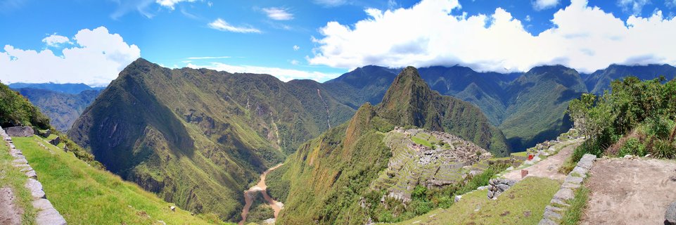 Panoramic view of Machu Picchu archaeological site with ancient stone ruins on green mountainside, Huayna Picchu peak in center, Urubamba River valley below, blue sky with white clouds