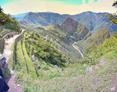 Panoramic view of Machu Picchu showing ancient stone terraces, tourists on pathways, Huayna Picchu mountain peak, and the Urubamba River winding through green valley