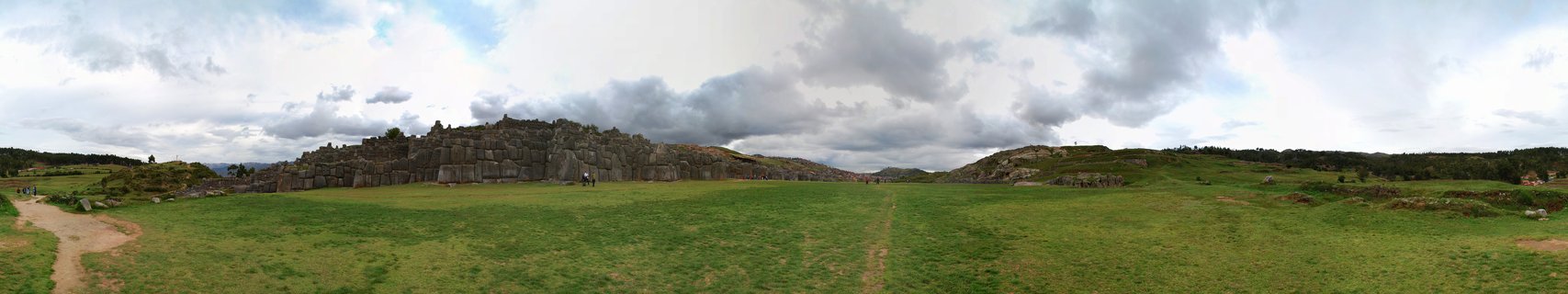 Wide panoramic photograph showing ancient stone ruins with large carved rock walls in the middle ground, green grassy field in foreground, cloudy sky above, Peru