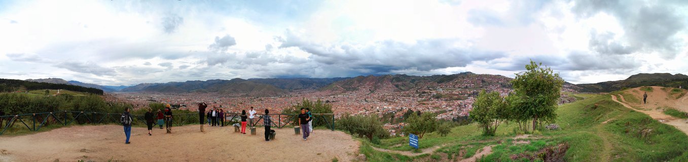 Panoramic view of Cusco city from a hillside viewpoint with tourists observing the red-roofed urban valley surrounded by Andean mountains under cloudy sky
