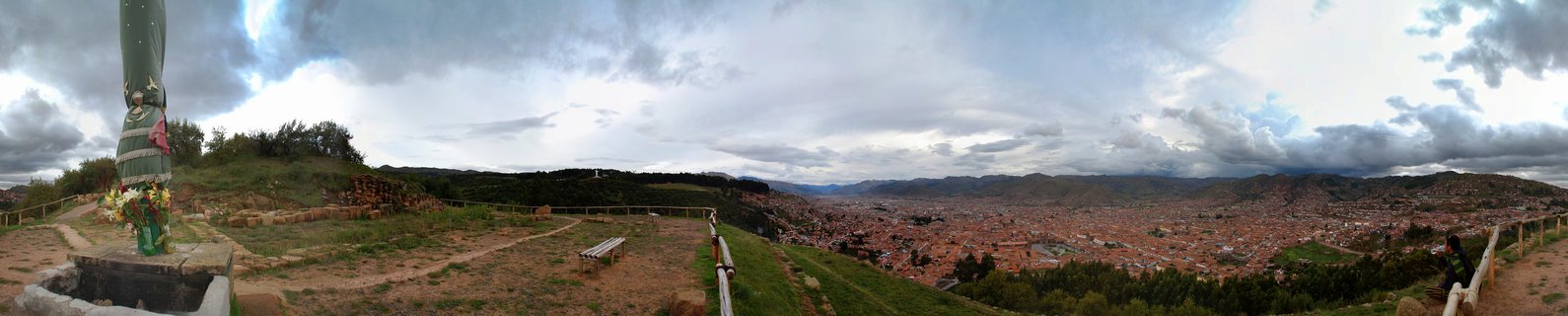 Panoramic landscape showing the city of Arequipa, Peru from an elevated viewpoint with a religious statue in the foreground, terracotta-roofed buildings filling the valley, and mountains under cloudy skies